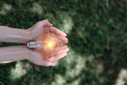 Hand hält leuchtende Glühbirne vor grünem Hintergrund - Symbol für energetische Zukunftsausrichtung